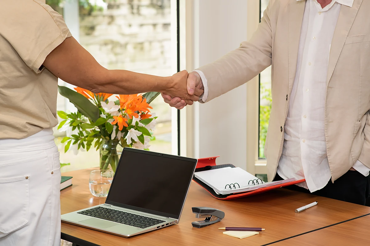 Aged Care Careers Melbourne: Two professionals, one in a tan t-shirt and the other in a light linen suit, shaking hands across a desk, symbolising a new BASSCARE aged care careers opportunity.