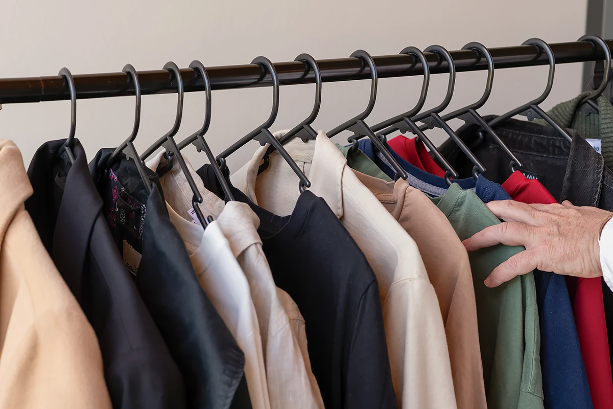 Ashburton Op Shop: A close-up shot of a hand browsing a densely packed rail of second-hand clothing in neutral and dark colours at The Ashy Op Shop, highlighting sustainable shopping.