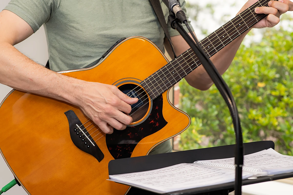 Residential Aged Care Ashburton: A close-up of a musician's hands playing an acoustic guitar during a music therapy session at Samarinda Lodge residential aged care, with sheet music on a stand and a green outdoor background.