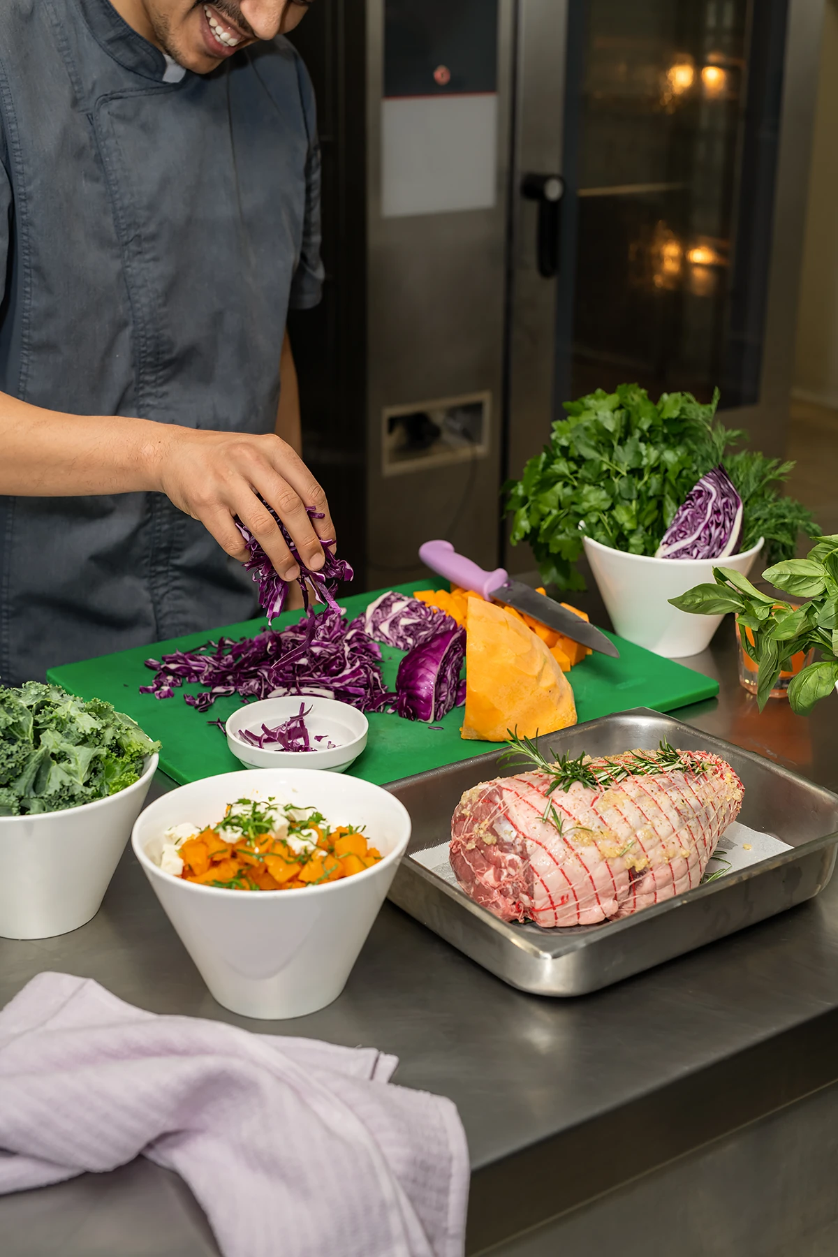 A BASSCARE Meals on Wheels chef, dressed in a grey uniform, smiling while preparing a meal, slicing fresh vegetables like red cabbage and sweet potato next to a tied lamb roast, in a commercial kitchen setting.