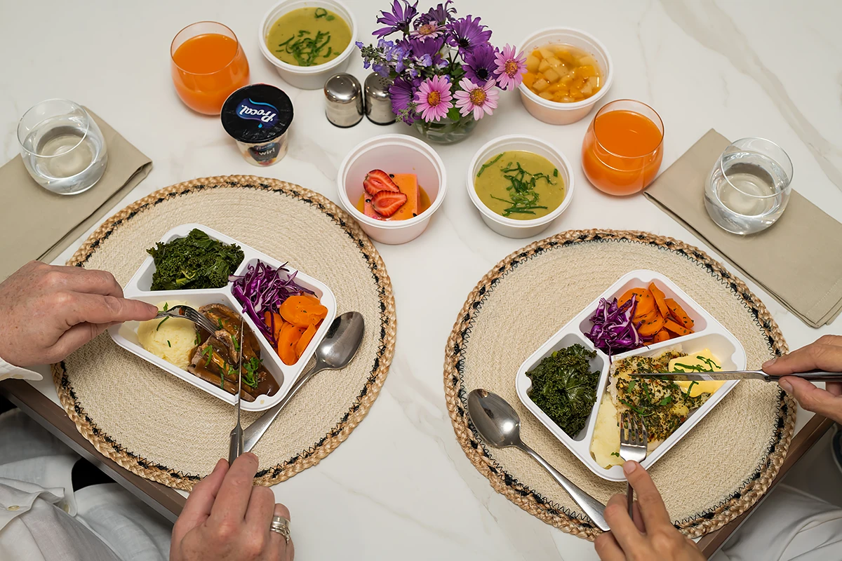 An overhead shot of two seniors eating their delivered Meals on Wheels meals at home from divided trays, including meat, mashed potato, vegetables, soup, juice and fruit.