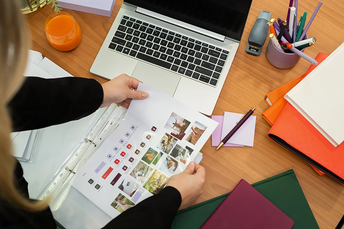 Volunteer with basscare: Overhead view of a volunteer's hands organising pages with colourful photo layouts and icons on a wooden desk with a laptop and notebooks, suggesting volunteer work for BASSCARE.