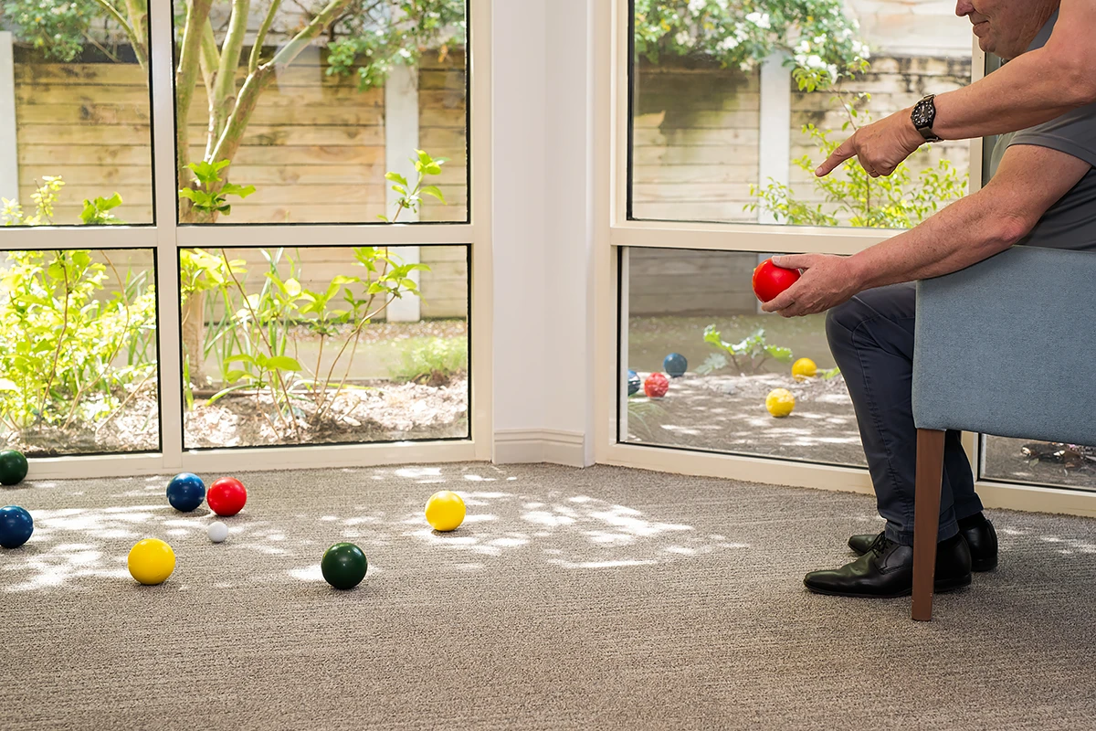 Volunteer with BASSCARE: A man sitting indoors, pointing toward a game of indoor bowls or bocce, with coloured balls scattered on the carpet and the garden visible through large window panes.